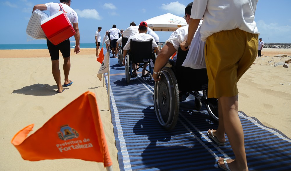 a imagem mostra pessoas de cadeiras de rodas sendo conduzidas pela rampa de acesso da praia acessível